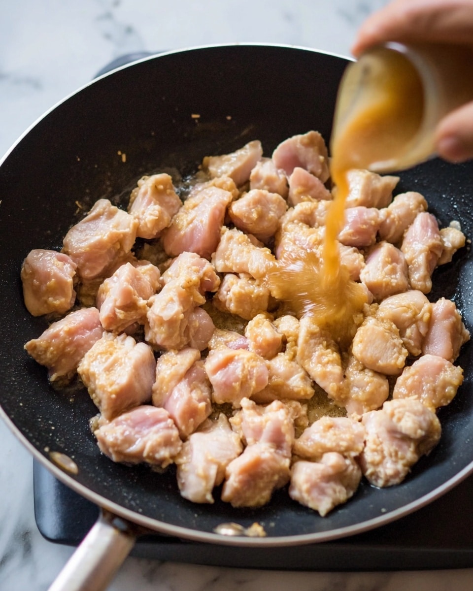A close-up view of small raw chicken pieces with a light beige and pink color arranged in a single layer inside a black frying pan, with a woman's hand pouring a light brown sauce over the chicken, creating a splash in the center. The pan sits on a white marbled surface. Photo taken with an iphone --ar 4:5 --v 7