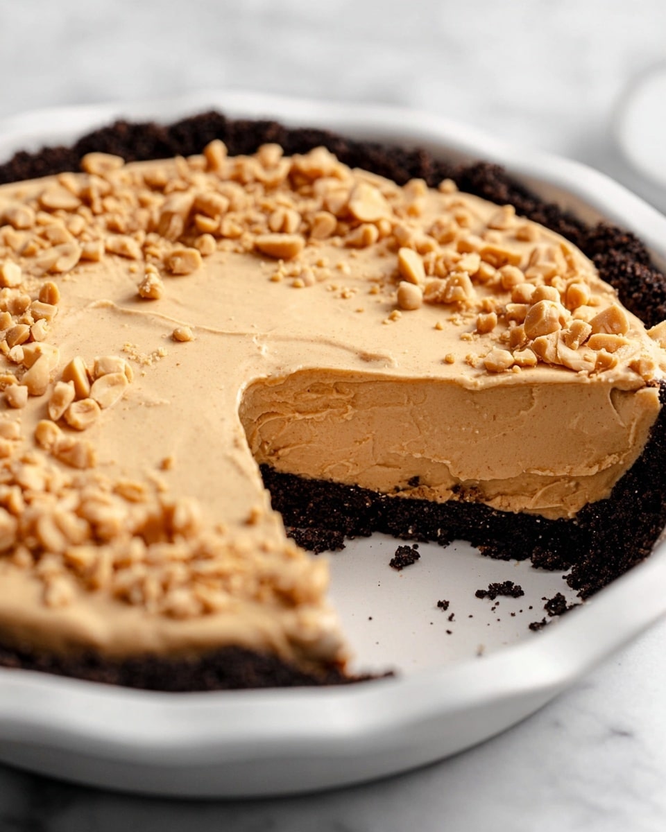 This image shows a two-layer peanut butter pie in a white pie dish on a white marbled surface. The bottom layer is a dark, crumbly chocolate crust forming a thick base and edges. Above it is a thick, smooth, and creamy peanut butter filling in a light tan color, evenly spread and slightly textured. The top surface is sprinkled with rough chopped peanuts, adding a crunchy texture contrast. The pie has a slice removed, clearly showing the height and thickness of each layer. Photo taken with an iphone --ar 4:5 --v 7