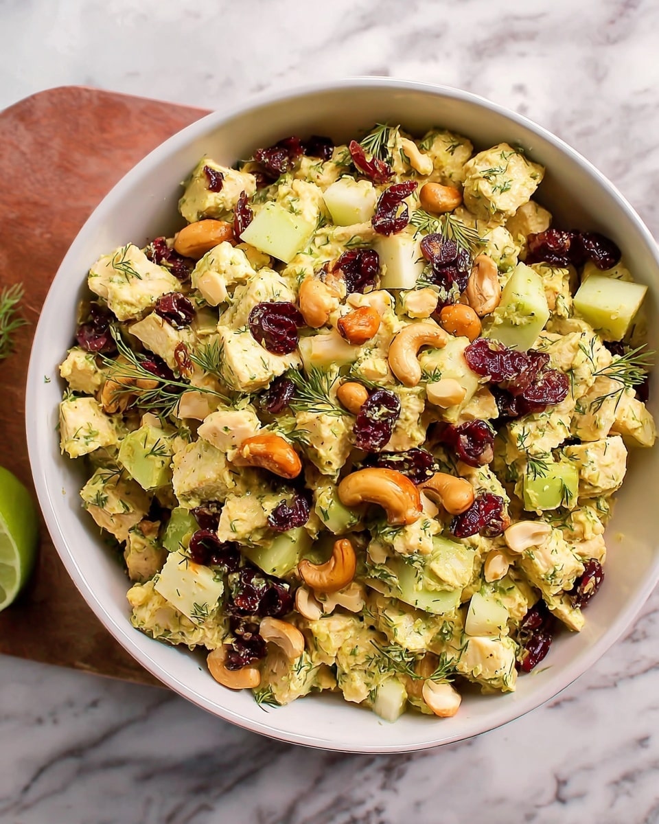 The image shows a white bowl filled with a colorful salad made of small, uneven cubes and pieces. The dish has three main layers: light yellow chicken pieces mixed with small chopped green apples and celery, which add fresh green tones. Scattered on top are dark red dried cranberries and light orange cashew nuts. Thin green herbs, possibly dill, are sprinkled throughout, adding detail and a fresh look. The bowl sits on a white marbled surface with a small piece of lime visible on the left side. The texture of the salad looks fresh and slightly creamy. photo taken with an iphone --ar 4:5 --v 7