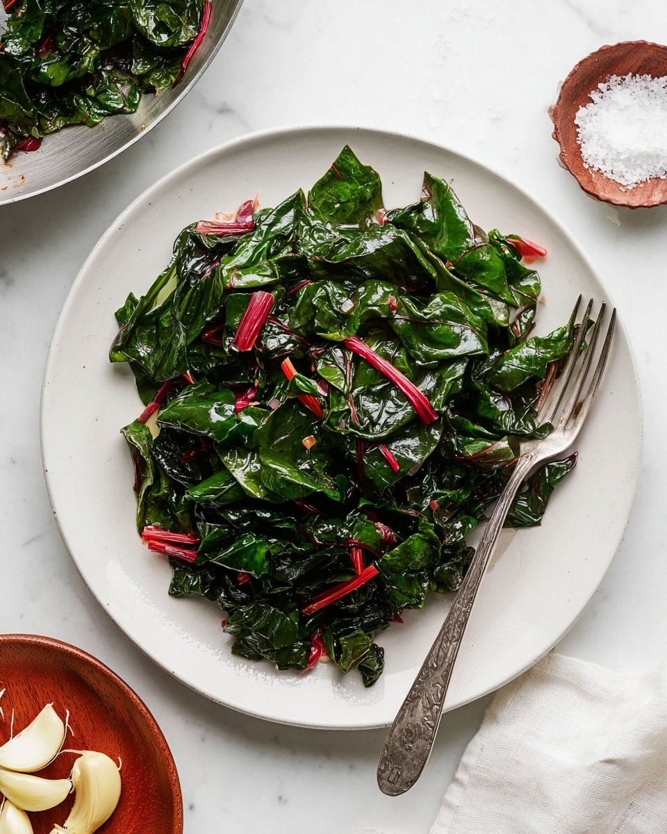 The image shows a white round plate filled with cooked leafy greens that are dark green with some bright red stems mixed in. The greens look shiny and soft, piled loosely in the center of the plate, with a silver fork resting on the right side. The plate is placed on a white marbled surface with a small round dish of coarse salt above it and a wooden plate with garlic cloves below. There is also a silver pan with more cooked greens visible on the left side of the image. photo taken with an iphone --ar 4:5 --v 7