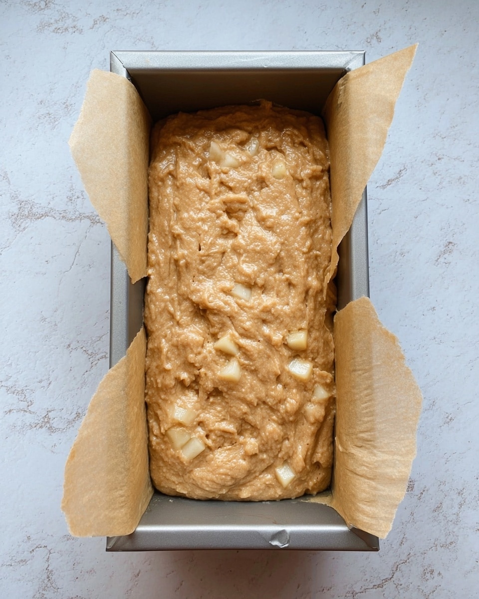 A rectangular metal baking pan lined with light brown parchment paper is filled with a thick, light brown batter. The batter contains visible small chunks of pale yellow fruit mixed evenly throughout the surface, giving it a textured look. The pan rests on a white marbled surface, showing a clean and simple background with soft light. The photo taken with an iphone --ar 4:5 --v 7