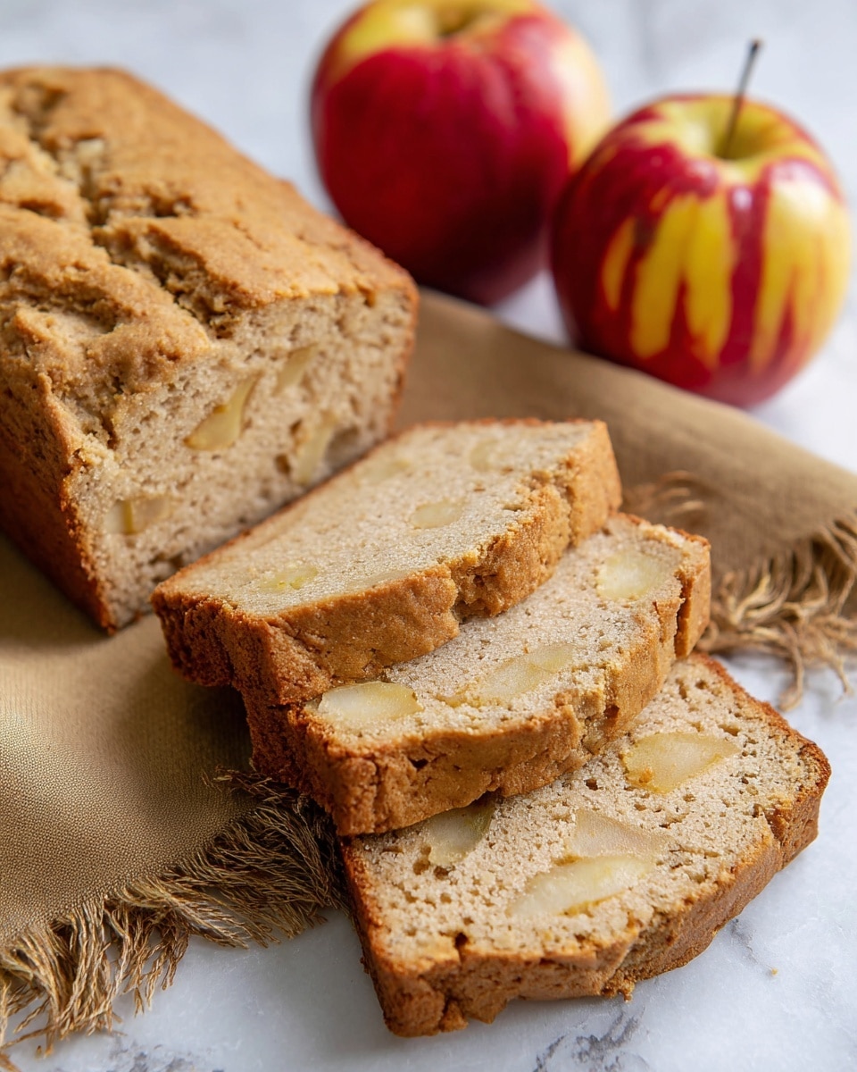 The image shows a loaf of apple bread with five visible slices laid out in front of the remaining loaf. The bread has a light brown color with soft, spongy texture and small, pale yellow pieces of apple scattered throughout each slice. The bread rests on a white marbled surface, partially covered by a brown fabric with fringed edges. In the background, there are two red apples with yellow streaks, positioned close together. The overall setting is bright and clean, highlighting the natural colors of the bread and apples. photo taken with an iphone --ar 4:5 --v 7