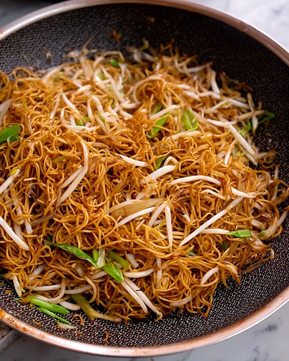 The image shows a pan filled with a single layer of golden-brown fried noodles mixed with white bean sprouts and green onion pieces. The noodles have a thin, slightly crispy texture, and the bean sprouts add a glossy, smooth contrast. The green onion pieces are bright and fresh, scattered evenly through the noodles. The pan has a dark textured, hexagonal pattern inside and a smooth metallic rim. The background features a white marbled surface. photo taken with an iphone --ar 4:5 --v 7