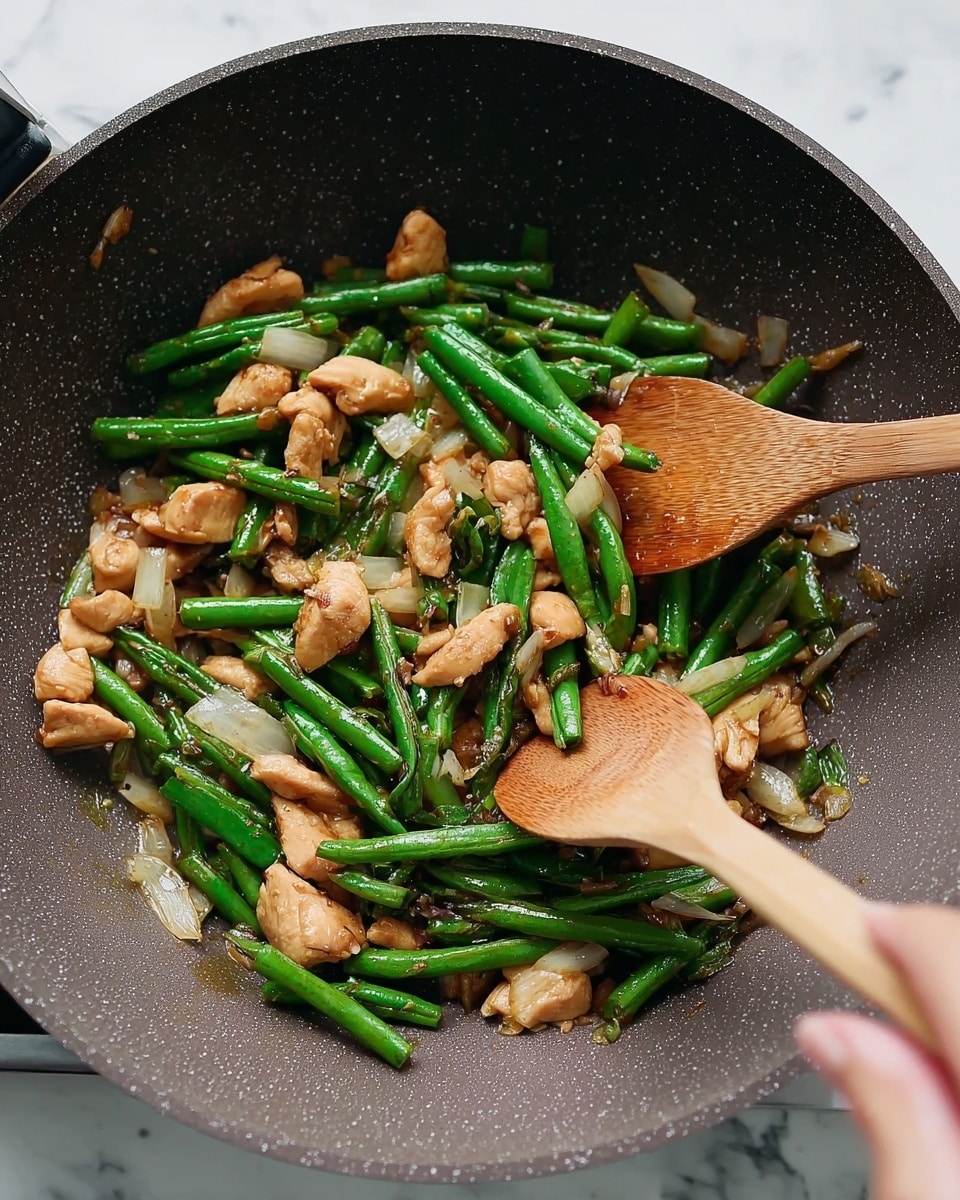 A dark gray wok filled with a stir-fry of three main layers: bright green chopped long beans, light brown pieces of cooked chicken scattered evenly, and translucent white chunks of onion mixed throughout. The green beans are fresh and firm, lying mostly on top and around the chicken. The chicken pieces are medium-sized with a shiny cooked surface, sitting mingled with the green beans and onions. The onions add a soft texture and light color contrast in the dish. Two wooden spoons are stirring the food, held by a woman's hand on the left side. The background surface is a white marbled texture. Photo taken with an iphone --ar 4:5 --v 7