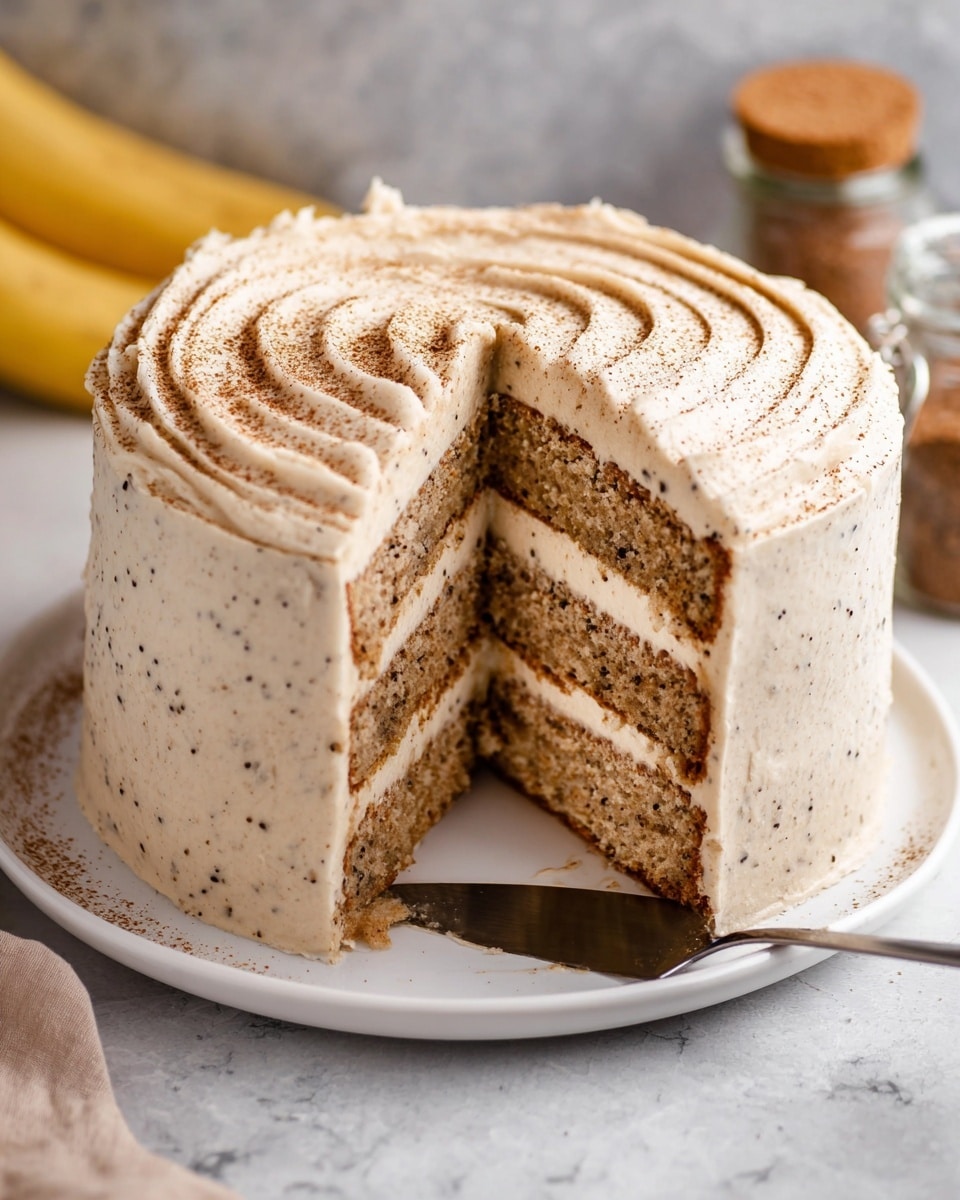 This image shows a tall three-layer round cake on a white plate placed on a white marbled surface. Each cake layer is light brown with dark small specks, separated and covered by smooth, creamy beige frosting. The cake is fully coated with the same frosting on all sides and top, with gentle swirls visible on the surface. A slice is cut out, showing the inner layers clearly, and the top edge of the cake has a light dusting of cocoa powder. A metal cake server rests under the cut slice, and there are blurred bananas and a small jar with a cork in the background. Photo taken with an iphone --ar 4:5 --v 7