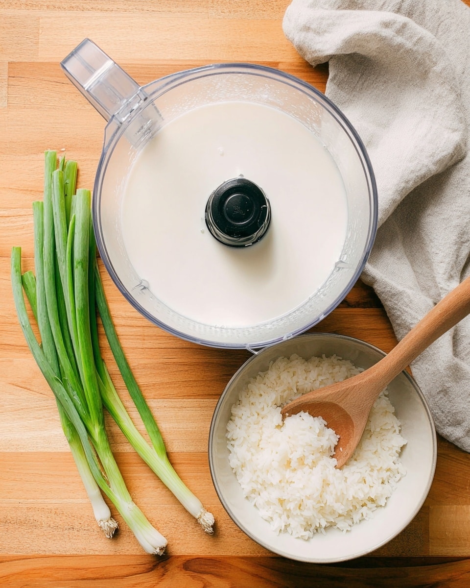 The image shows a top view of a food processor containing a smooth white liquid filling about two-thirds of its clear container, placed on a wooden surface. To the right, there is a white bowl filled halfway with cooked white rice, and a wooden spoon resting inside the bowl with some rice on it. To the left of the food processor, there are five pieces of fresh green onions with vivid green stalks laying on the wooden surface. A light gray cloth is casually placed in the upper right corner of the image. photo taken with an iphone --ar 4:5 --v 7