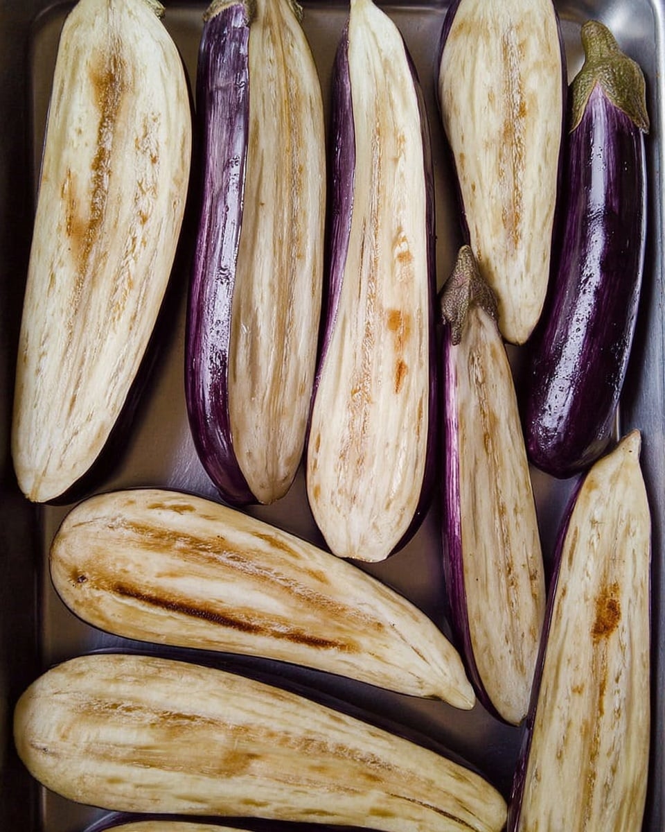 The image shows several long, halved eggplants laid flat on a metal tray. Each eggplant is cut lengthwise, revealing a pale cream inside with a soft, slightly spongy texture, and a dark purple shiny skin on the outside edges. The inside layers have a few small seeds scattered throughout, and some of the flesh has light brown marks from roasting or grilling. The eggplants are arranged side by side, filling the entire tray with their curved shapes and smooth surfaces. photo taken with an iphone --ar 4:5 --v 7