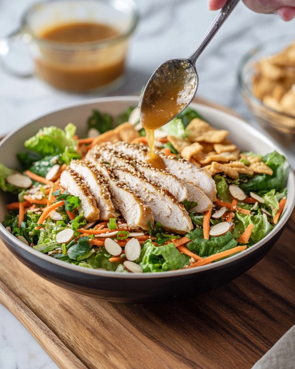 A salad in a white bowl with a dark outer rim sits on a wooden board against a white marbled background. The base layer is bright green leafy lettuce covered with thin orange carrot strips and sliced light brown almonds. Over this, there are light beige fried wonton strips and finely chopped green onions. On the top is a layer of sliced light golden grilled chicken, sprinkled with sesame seeds. A woman's hand holds a spoon pouring a golden brown dressing over the chicken. In the background, there is a glass cup filled with more dressing. photo taken with an iphone --ar 4:5 --v 7