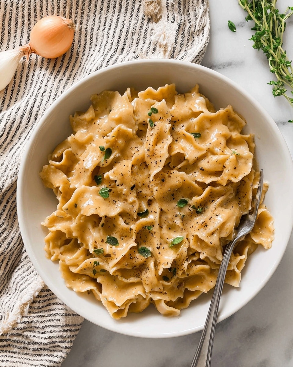 A white bowl is filled with about two layers of wavy, wide pasta covered in creamy, light brown sauce. The pasta has a smooth, soft texture with ruffled edges, and small green herb leaves are scattered on top along with a light sprinkle of black pepper. A silver fork rests inside the bowl on the right side, partially submerged in the pasta. The bowl is placed on a white marbled surface, with a striped cloth napkin in the upper left corner and a whole light yellow onion near the bottom left, with some green herb sprigs in the upper right corner. Photo taken with an iphone --ar 4:5 --v 7