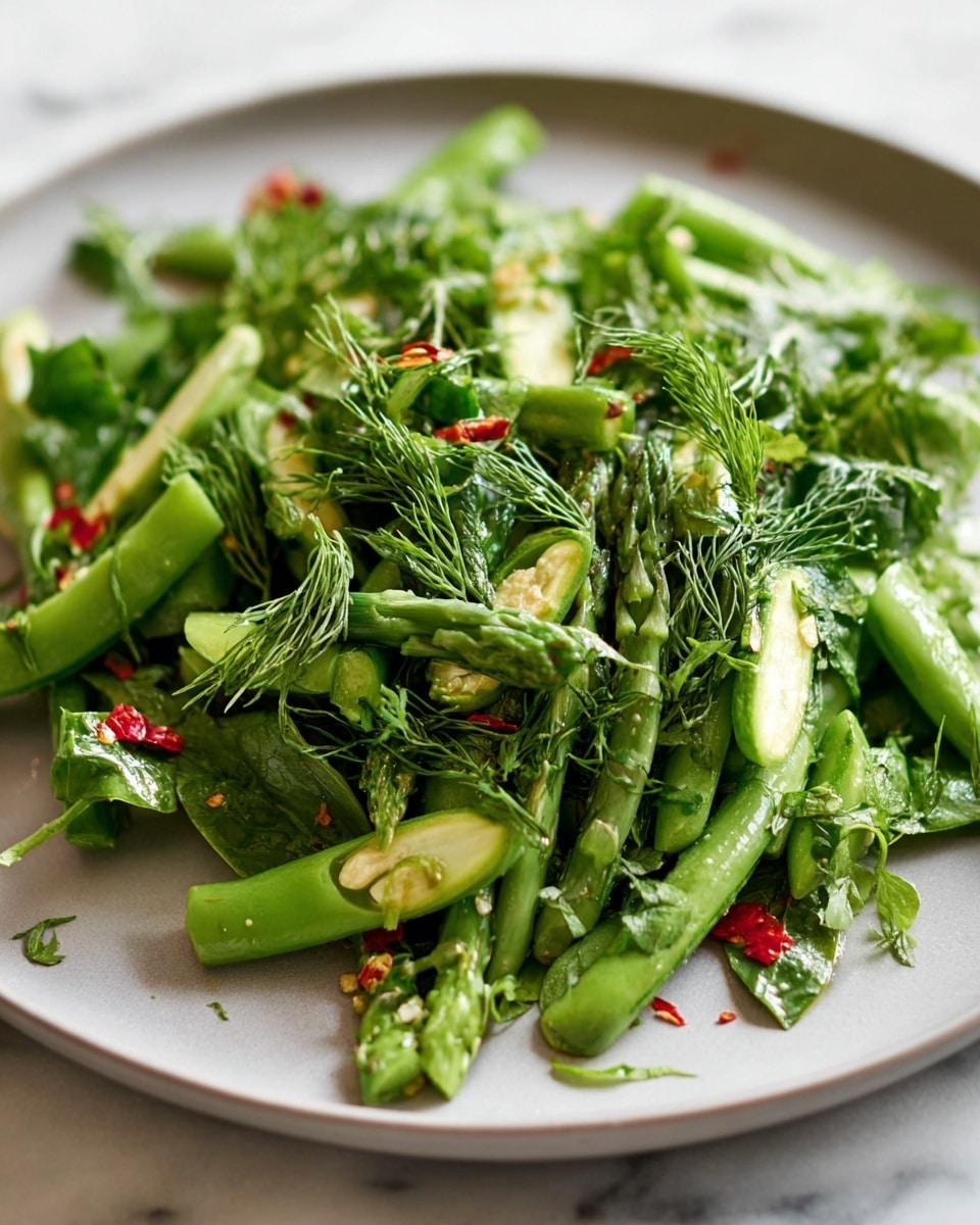 The image shows a close-up of a white plate filled with a fresh green vegetable salad. The dish has a base layer of light green leafy herbs, topped with thicker stalks of bright green asparagus and snap peas cut into pieces. Over this, there is a scattering of fine, delicate dill fronds adding texture and a few small red chili flakes for a touch of color. The plate rests on a white marbled surface. photo taken with an iphone --ar 4:5 --v 7